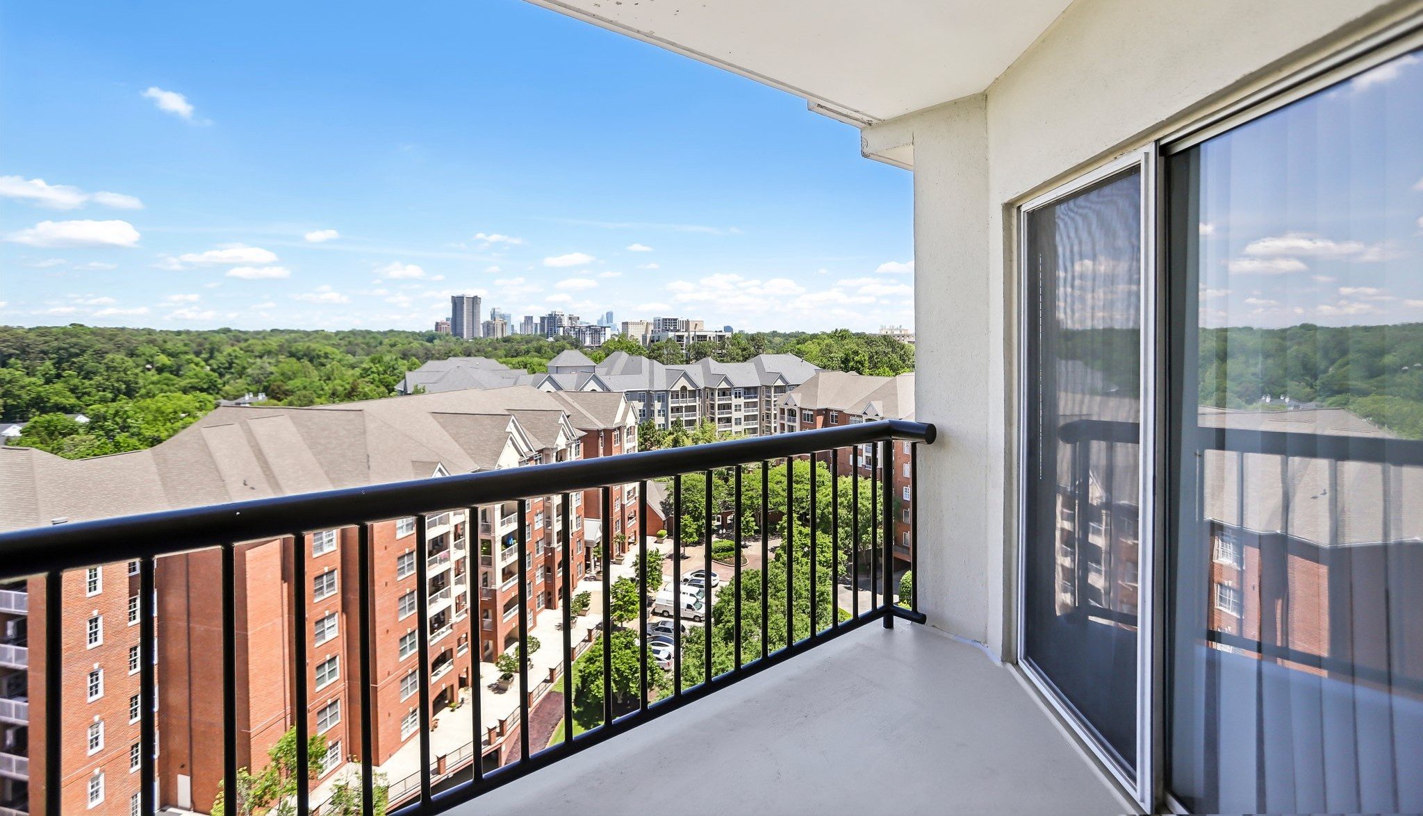A balcony overlooking apartment rooftops and a distant skyline, showing a black railing and a glass sliding door.